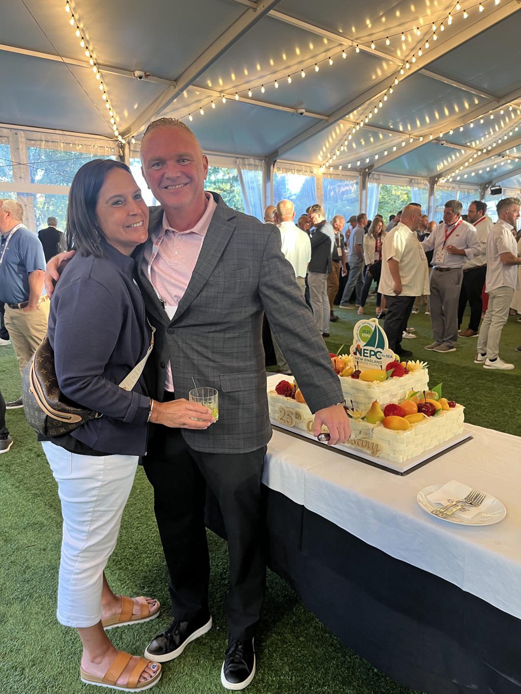 A couple posing by a decorated cake at a festive outdoor event under string lights.